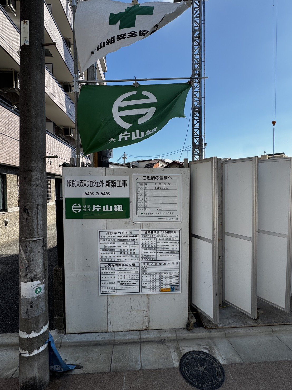 株式会社片山組の旗と仮囲い、各種許可票が掲示された現場入口